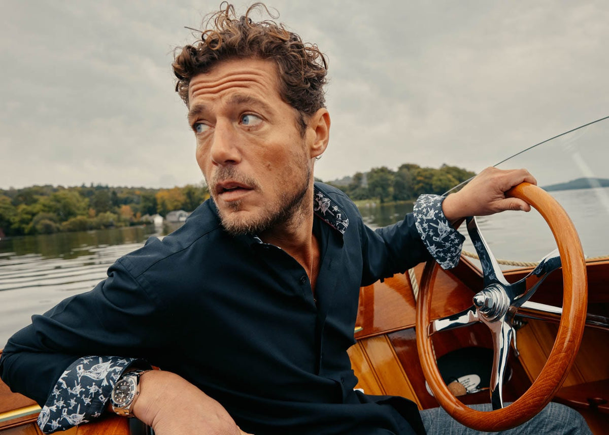 Man wearing a dark shirt with patterned cuffs at a boat’s wheel